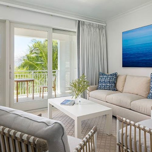 A bright, modern living room with white furniture, blue accents, and large glass doors leading to a balcony with a view of tropical greenery and the ocean.