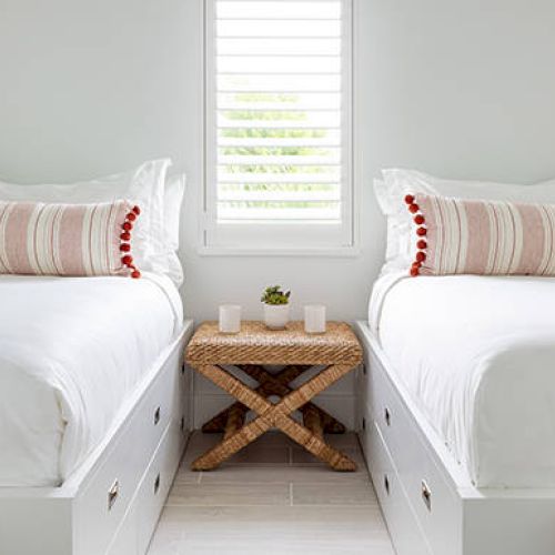 A bright bedroom with two white beds featuring striped pillows and a wicker table between them, under a window with shutters.