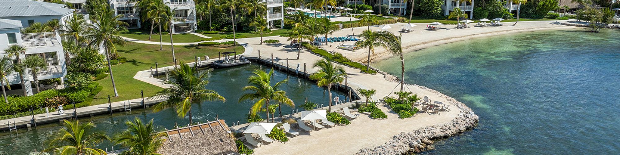 A picturesque beachfront resort with white buildings, lush greenery, palm trees, a sandy beach, and clear, calm ocean water covering rocks.