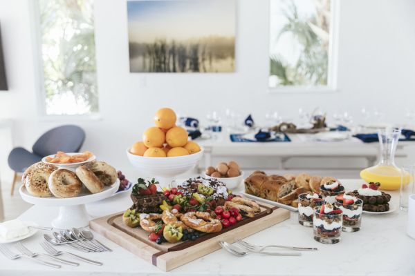 A table displaying a variety of breakfast foods, including bagels, fruit parfaits, pastries, fresh fruit, and juice, set in a bright dining room.