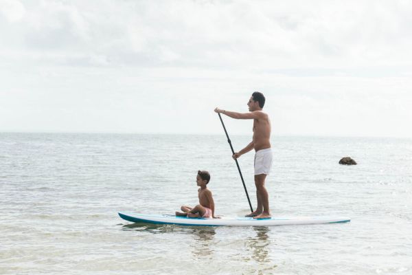 A person is stand-up paddleboarding with a child sitting in front on a calm ocean water. They both appear to be enjoying a leisurely time.