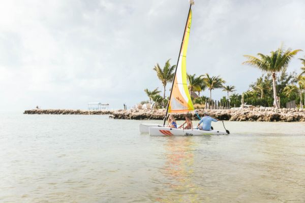 A small sailboat with three people on board is sailing near a tropical shoreline with palm trees and rocks in the background.