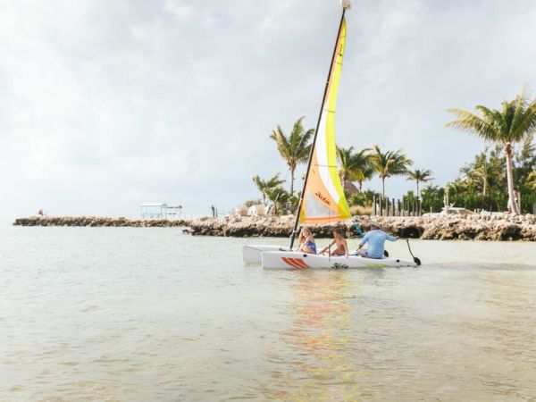 A small sailboat with three people on board is sailing near a tropical shoreline with palm trees and rocks in the background.