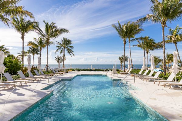 A luxurious pool area with sun loungers and palm trees, overlooking a serene ocean view under a clear blue sky with some wispy clouds.