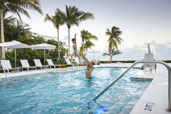 A person lifts a child in a swimming pool surrounded by lounge chairs and palm trees, under a clear sky.