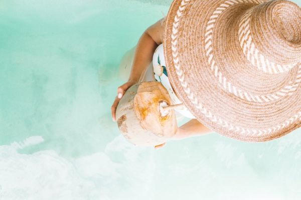 A person wearing a wide-brimmed hat stands or floats in clear water while holding and drinking from a coconut.