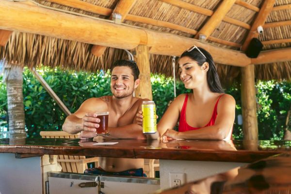 A man and woman sit at a tiki bar, smiling and enjoying drinks with a wooden roof and lush green background.