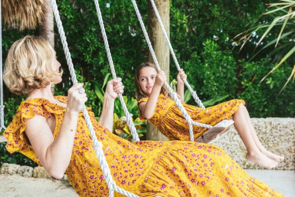 Two people in yellow dresses are sitting on swings under a rustic pergola, surrounded by lush greenery.