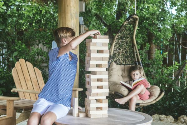 A person plays Jenga on a table outdoors, while another sits in a hanging chair. A bag is on the ground.