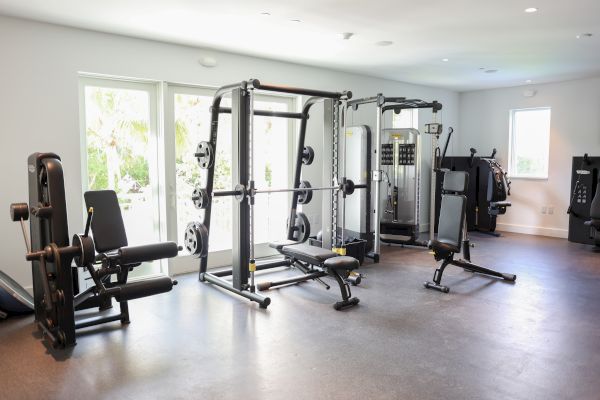 A home gym with various exercise equipment, including weight machines, benches, and a Smith machine, in a bright room with windows.