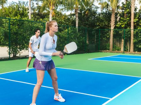 Two people are playing pickleball on an outdoor court surrounded by trees. They are both ready to hit the ball, with paddles in hand.