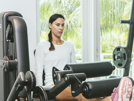 A person is using a leg extension machine in a gym, wearing a white top and pink shoes. The background has windows showing greenery outside.