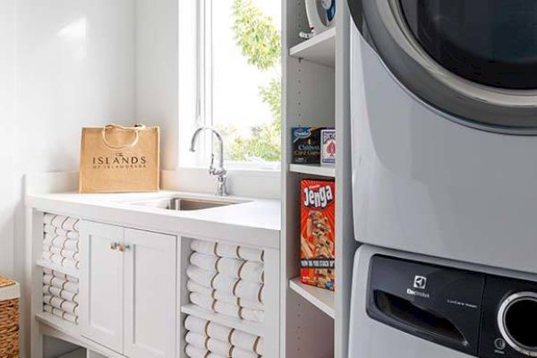 A laundry room with a sink, stacked washer/dryer, cabinet with neatly rolled towels, and shelves with board games and detergent.