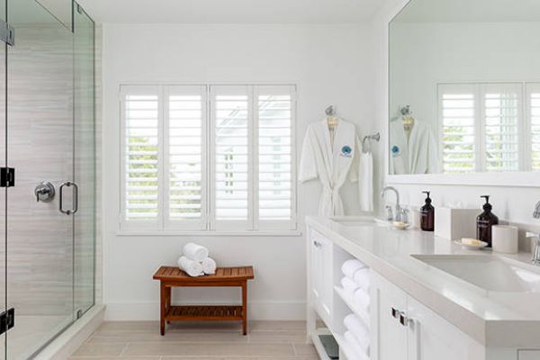 A modern bathroom with a glass-enclosed shower, double sink counter, robes on hooks, and folded towels on a wooden bench by a window.