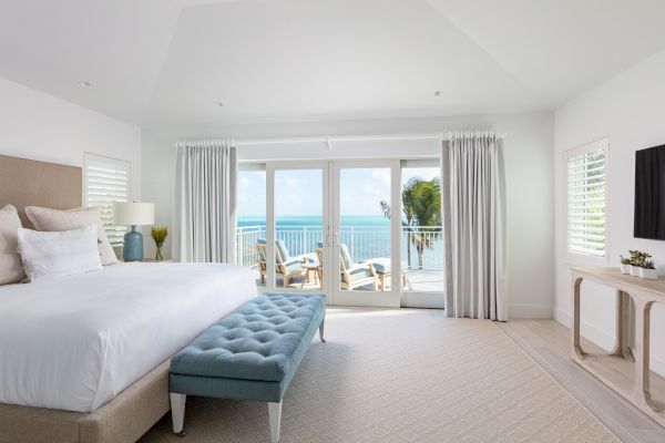 A bright bedroom with a large bed, a blue bench, a TV, and glass doors leading to a balcony with ocean views and outdoor seating, all in a serene setting.