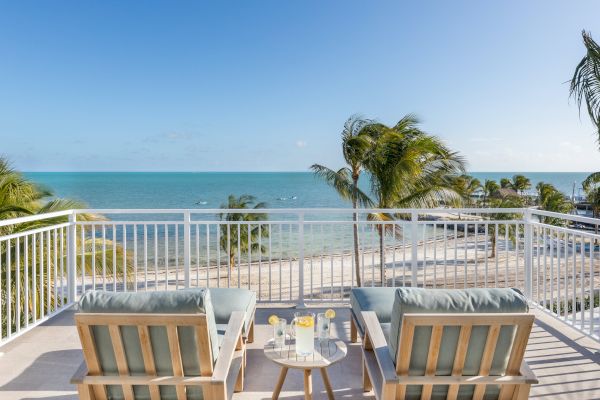 A scenic oceanview balcony with two lounge chairs, a table with drinks, and palm trees, overlooking a sandy beach under a clear blue sky.