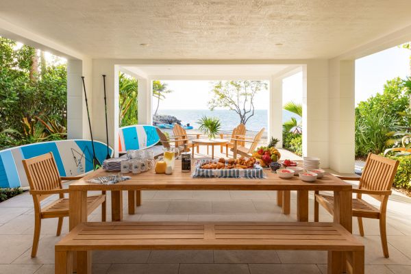 A patio setup with wooden tables and benches, a variety of breakfast foods, surfboards on the side, and a view of the ocean beyond.