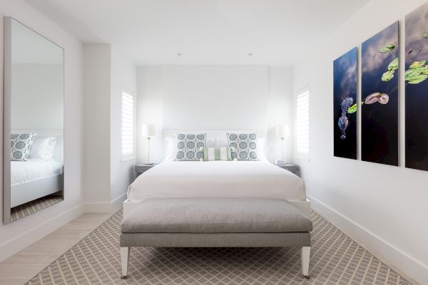 A modern bedroom with a bed, a bench at the foot, two side tables with lamps, a large mirror, and wall art featuring lily pads and flowers.