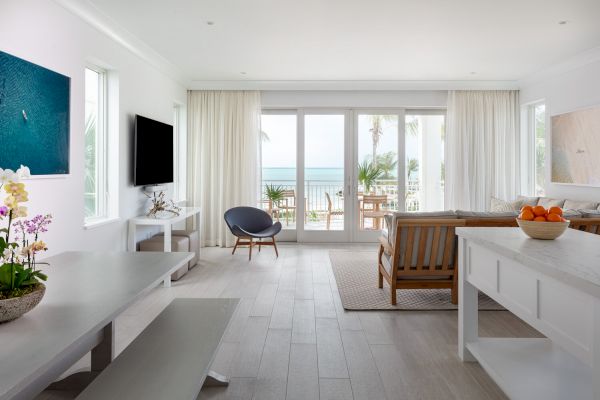 A bright, modern living room with white walls and furnishings, overlooking a balcony with a beach view, featuring a TV, sofa, and dining area.