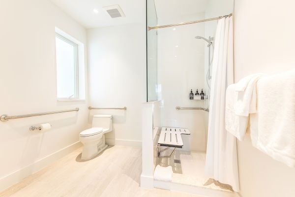 A clean, white bathroom featuring a toilet, shower area with a bench and grab bars, a towel rack with towels, and a window for natural light.