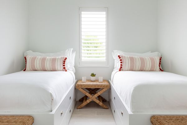 The image shows a minimalist bedroom with two twin beds, white linens, decorative pillows, a small wooden table between them, and a window with shutters.