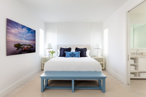 A well-lit bedroom with a bed, blue bench, white bedding, and two nightstands with lamps. A colorful painting adorns the wall. A bathroom is adjacent.