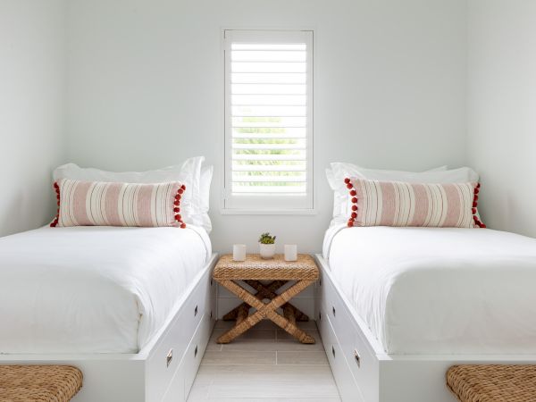 A minimalist bedroom with two white twin beds, a small window with shutters, a woven nightstand, and matching striped pillows and throws.