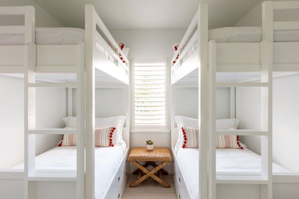 The image shows two sets of white bunk beds with striped pillows, a window with blinds in between, and a small wooden stool at the center.