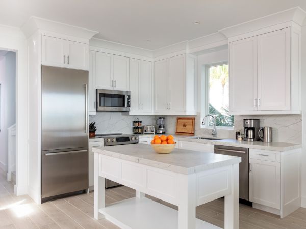 A modern kitchen with white cabinets, a stainless steel refrigerator, a microwave, and an island with a bowl of oranges on top.