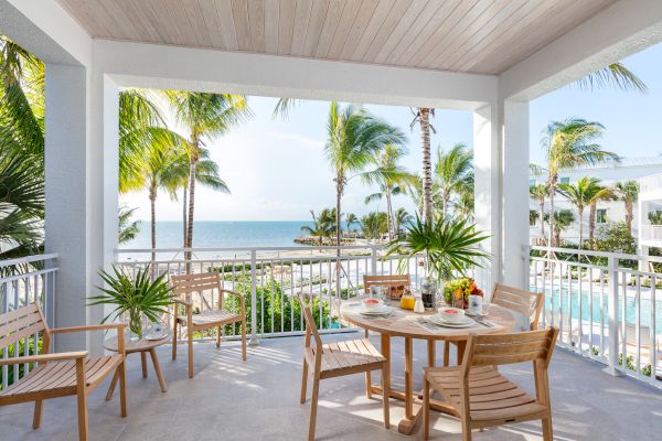 A patio with wooden furniture, breakfast items on the table, overlooking a pool, palm trees, and a beach with a clear blue sky in the background.