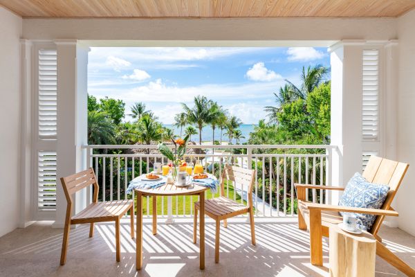 A balcony with a wooden table set for breakfast, overlooking a lush garden and the ocean, framed by palm trees and a bright, sunny sky.