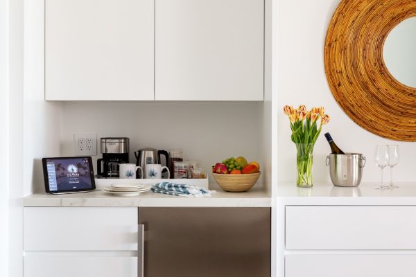 A modern kitchen corner with white cabinets, a stainless-steel fridge, a countertop with kitchen gadgets, flowers, a fruit bowl, and a round mirror.