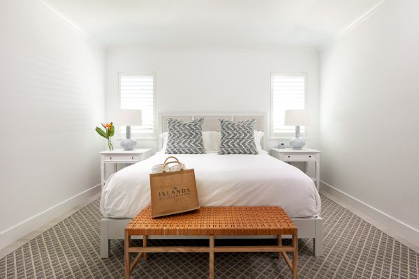 A neatly arranged bedroom with a large bed, two nightstands with lamps, a bag on the bed, a bench at the foot, and a vase with flowers.