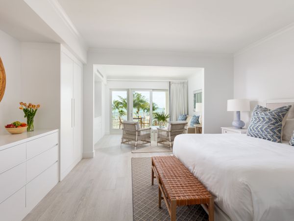 A bright, modern bedroom with a large bed, seating area, chest of drawers, and balcony view with palm trees.