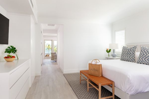 A modern, well-lit bedroom with white decor, a bed, a wicker bench, a large TV, and a view of a balcony with outdoor furniture in the distance.