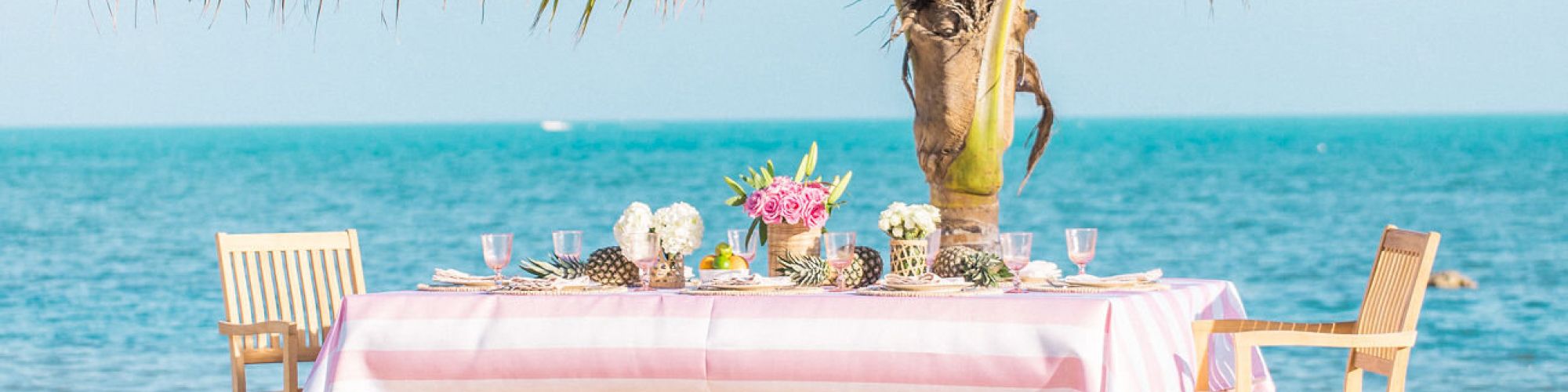 A beachside table with a pink tablecloth is set with flowers and dishes, shaded by a palm tree, overlooking the ocean.