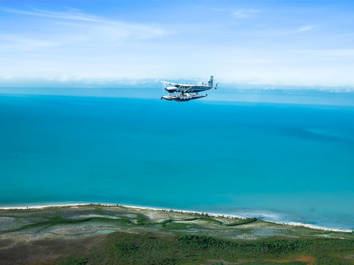 A seaplane flies over a vibrant blue body of water near a green coastline under a clear sky, capturing a serene and picturesque aerial view.