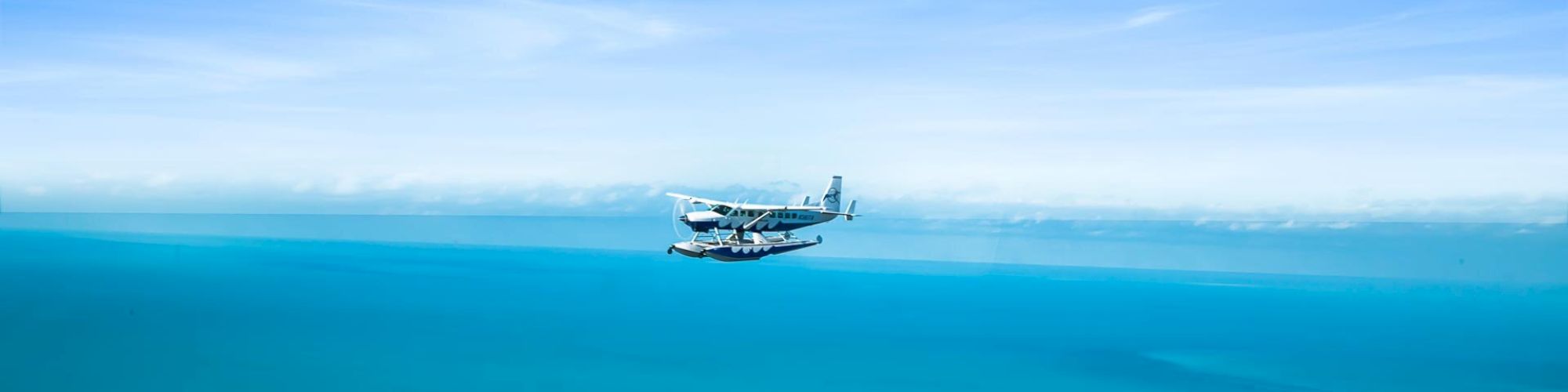 A small seaplane flies over a vast turquoise ocean with a green coastline visible below. The sky above is clear with some light clouds on the horizon.