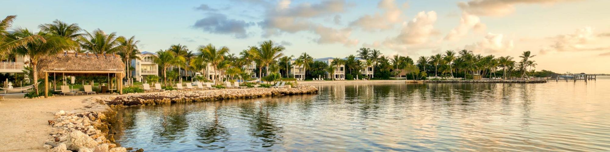 The image shows a scenic coastal view with a rocky shoreline, palm trees, and houses in the background under a blue sky with clouds at sunset.