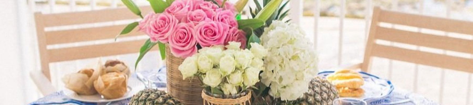 A table by the sea is set for a meal with plates of food, glasses of juice, and floral centerpieces, creating a picturesque, beachside dining scene.