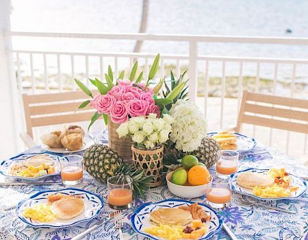 A table by the sea is set for a meal with plates of food, glasses of juice, and floral centerpieces, creating a picturesque, beachside dining scene.