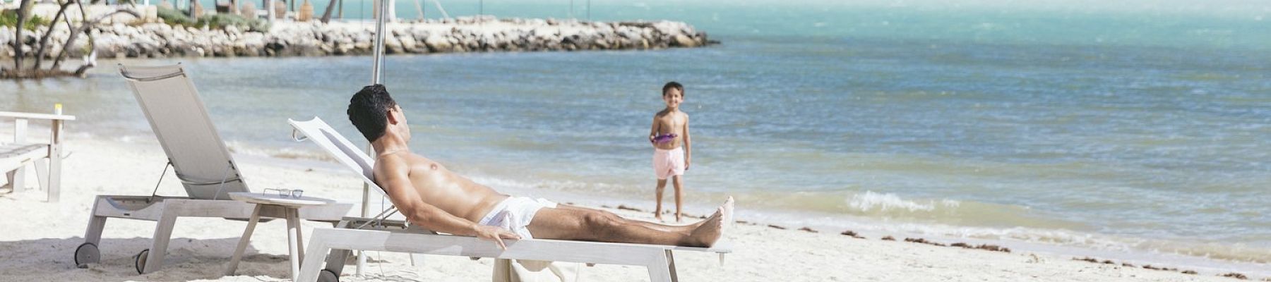 A man lounges on a beach chair under an umbrella, while a child stands nearby on sandy beach by turquoise water and palm trees in the background.