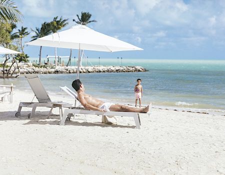 A man lounges on a beach chair under an umbrella, while a child stands nearby on sandy beach by turquoise water and palm trees in the background.