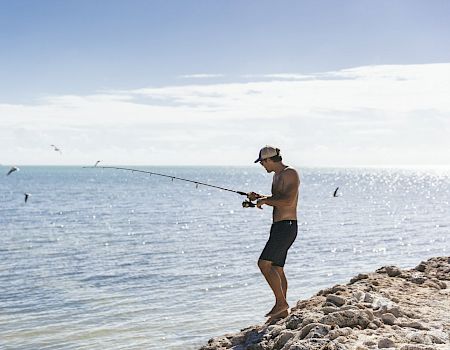 A person is fishing while standing on rocky terrain beside a body of water, with seagulls flying in the background under a clear sky.