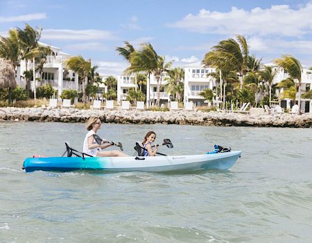 Two people are kayaking on a body of water with white buildings and palm trees in the background under a blue sky with scattered clouds.