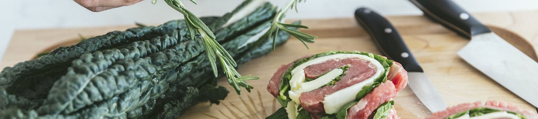 A person prepares a meal with kale and rolled meat, garnishing it with herbs on a wooden cutting board, alongside two knives.