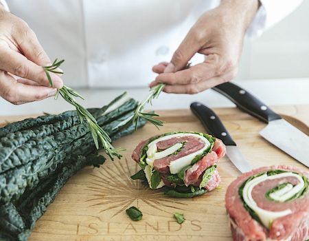 A person prepares a meal with kale and rolled meat, garnishing it with herbs on a wooden cutting board, alongside two knives.