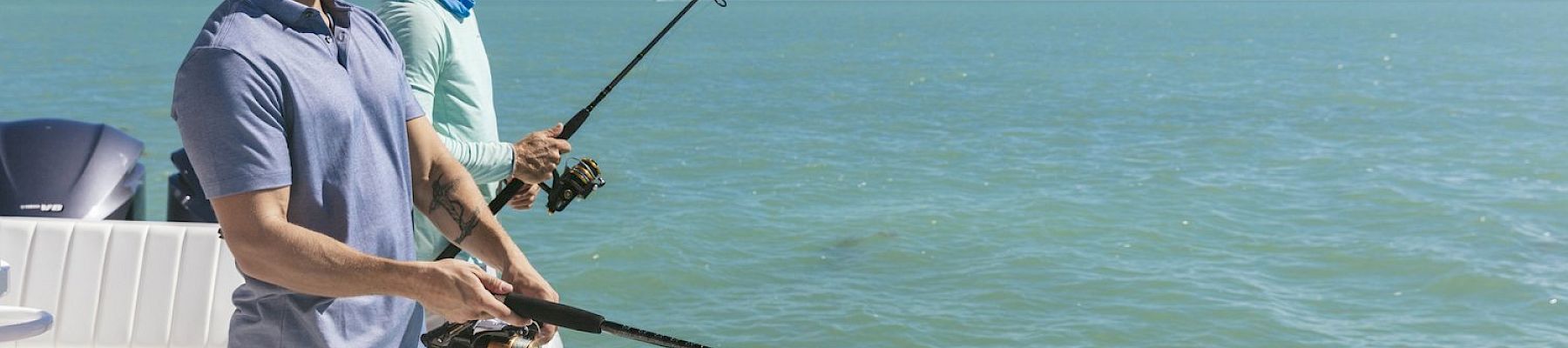 Two men are standing on a boat, both holding fishing rods and fishing in the calm, expansive ocean under a clear blue sky.