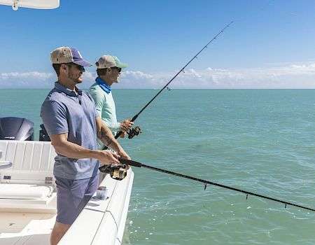 Two men are standing on a boat, both holding fishing rods and fishing in the calm, expansive ocean under a clear blue sky.