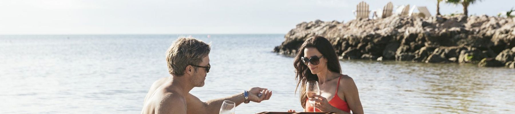 A man and a woman are playing backgammon on a table in shallow ocean water, both holding wine glasses, with a palm tree in the background.